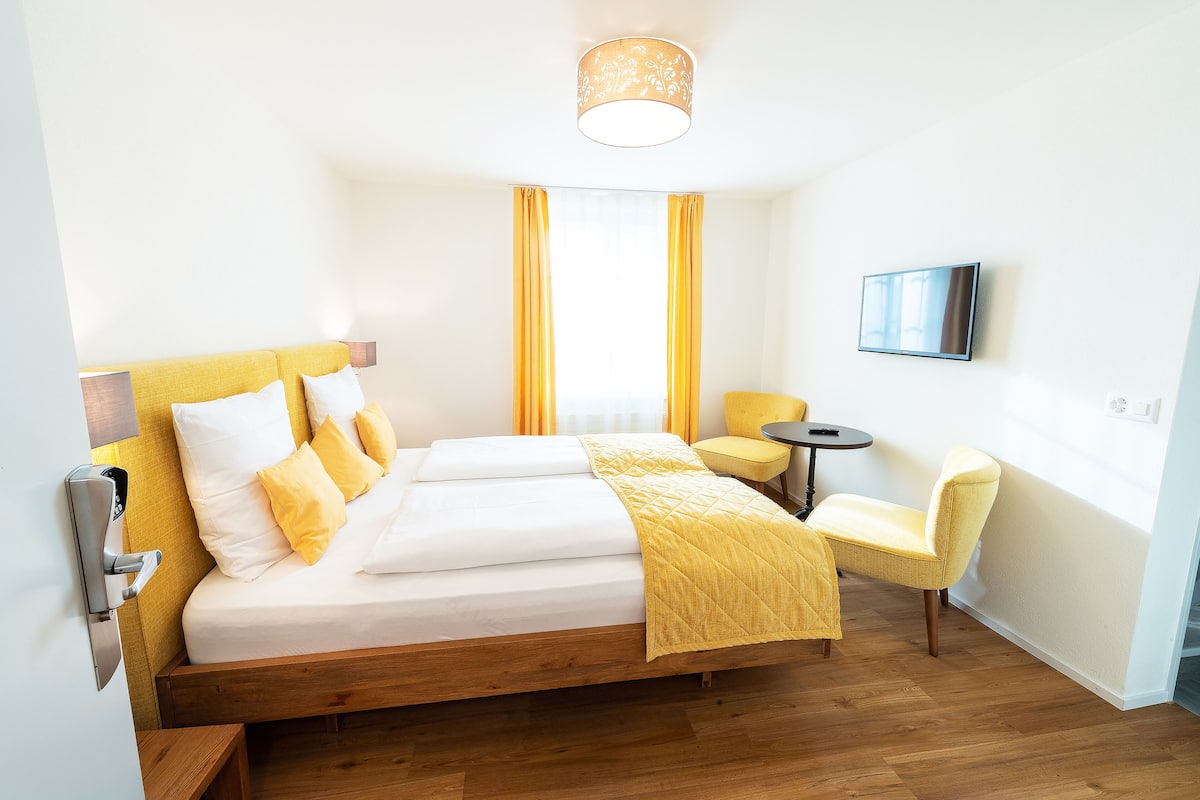 Habitación de hotel con una cama de madera, ropa de cama amarilla, una pequeña mesa redonda y una silla.