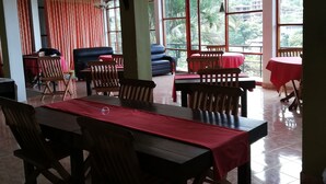 A dining area with wooden tables and chairs, a red tablecloth, and large windows with a view of greenery. - Tea Garden Holiday Inn (Ella)