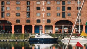 A brick building with the letters "TATE" on the facade, a calm water body with boats, and a clear sky. - The Liverpool Pod - Hostel (Liverpool)