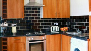 A compact kitchen with wooden cabinets, a built-in oven, a washing machine, and a tiled backsplash. - The Liverpool Pod - Hostel (Liverpool)