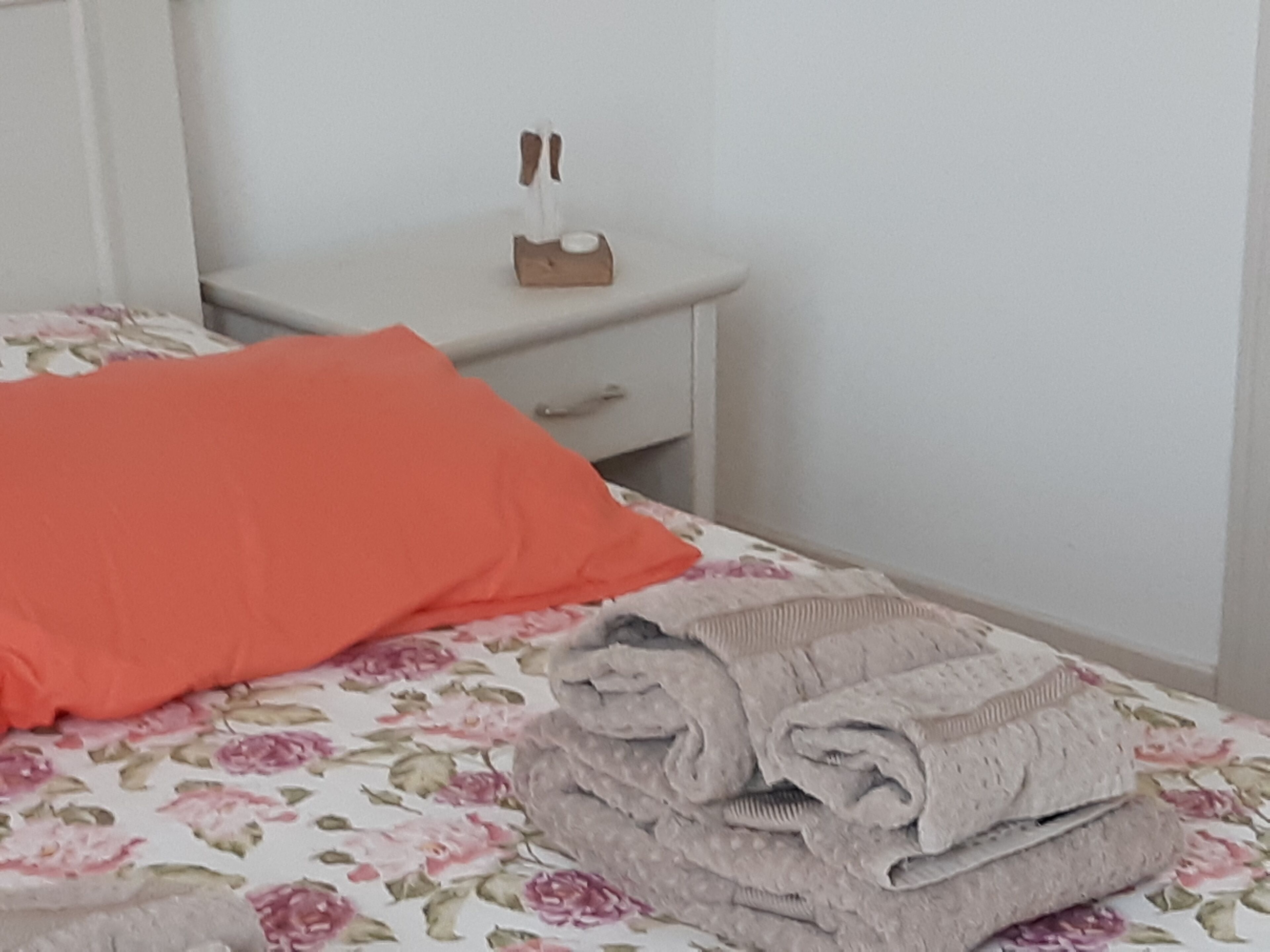 A neatly made bed with a floral bedspread, an orange pillow, and folded towels.