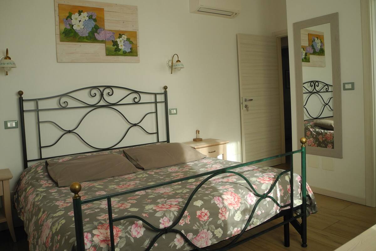Un dormitorio con cama, mesita de noche, espejo y un cuadro floral en la pared.
