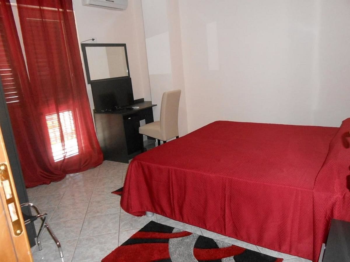 A bedroom with a bed, a desk, a chair, and a window with red curtains.