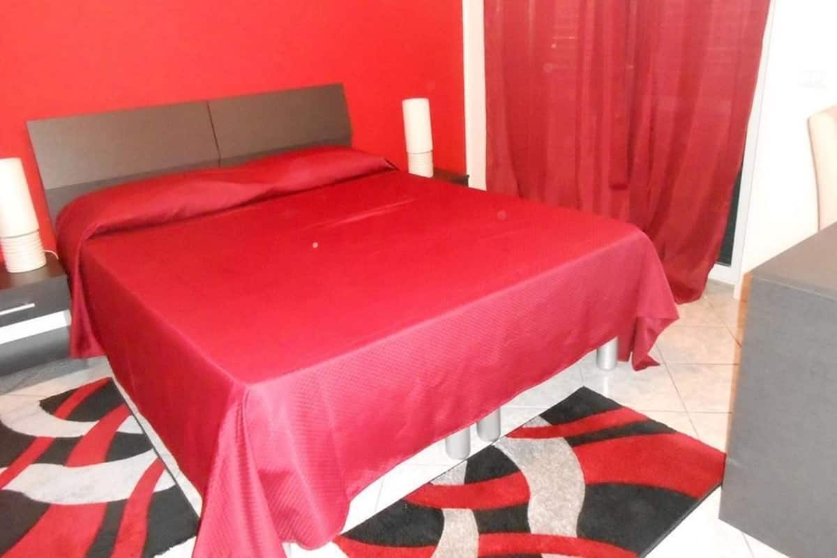Uma cama com um cobreleto vermelho, cabeceira cinza e tapete estampado.
