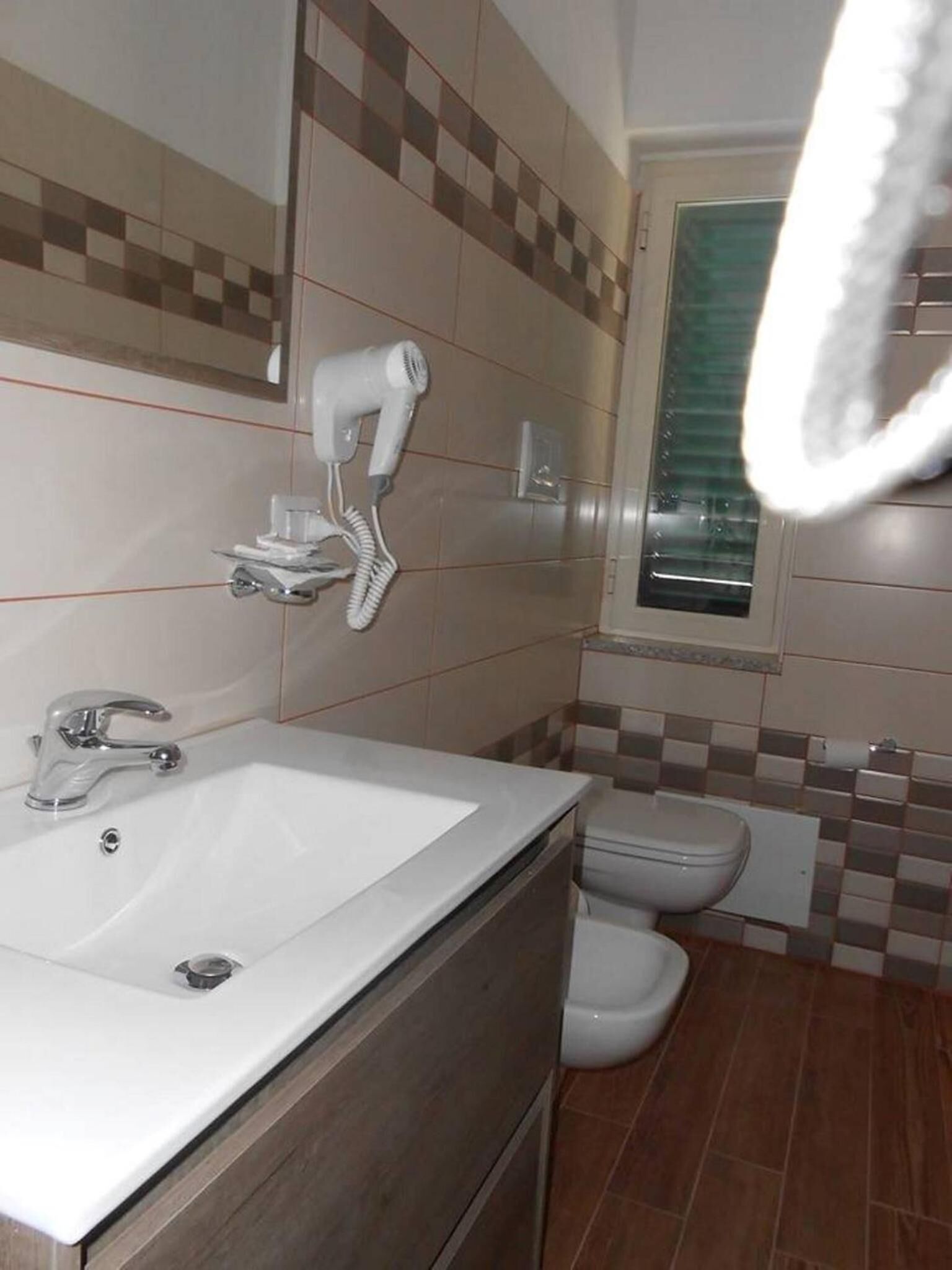Une salle de bain avec un lavabo blanc, un sèche-cheveux, des toilettes et une fenêtre avec des stores.