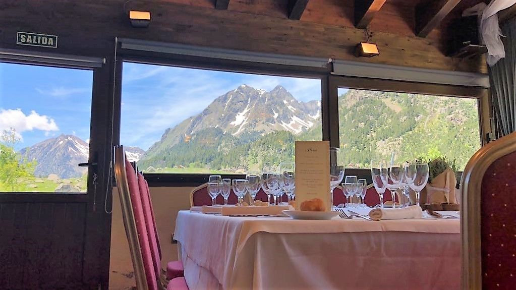 Un comedor con una mesa lista para una comida, un atril para el menú y vistas a montañas a través de amplias ventanas.