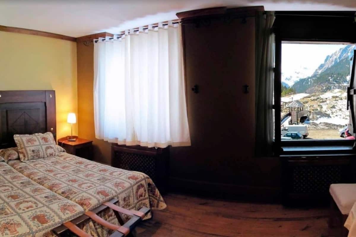 A bedroom with a bed, a window with curtains, a nightstand, and a view of mountains.