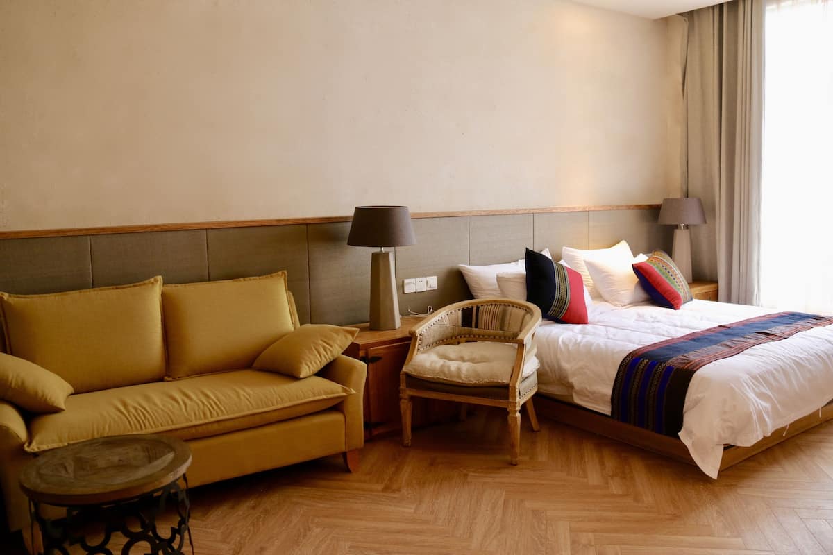A hotel room with a sofa, a chair, a bed with pillows, and a lamp.