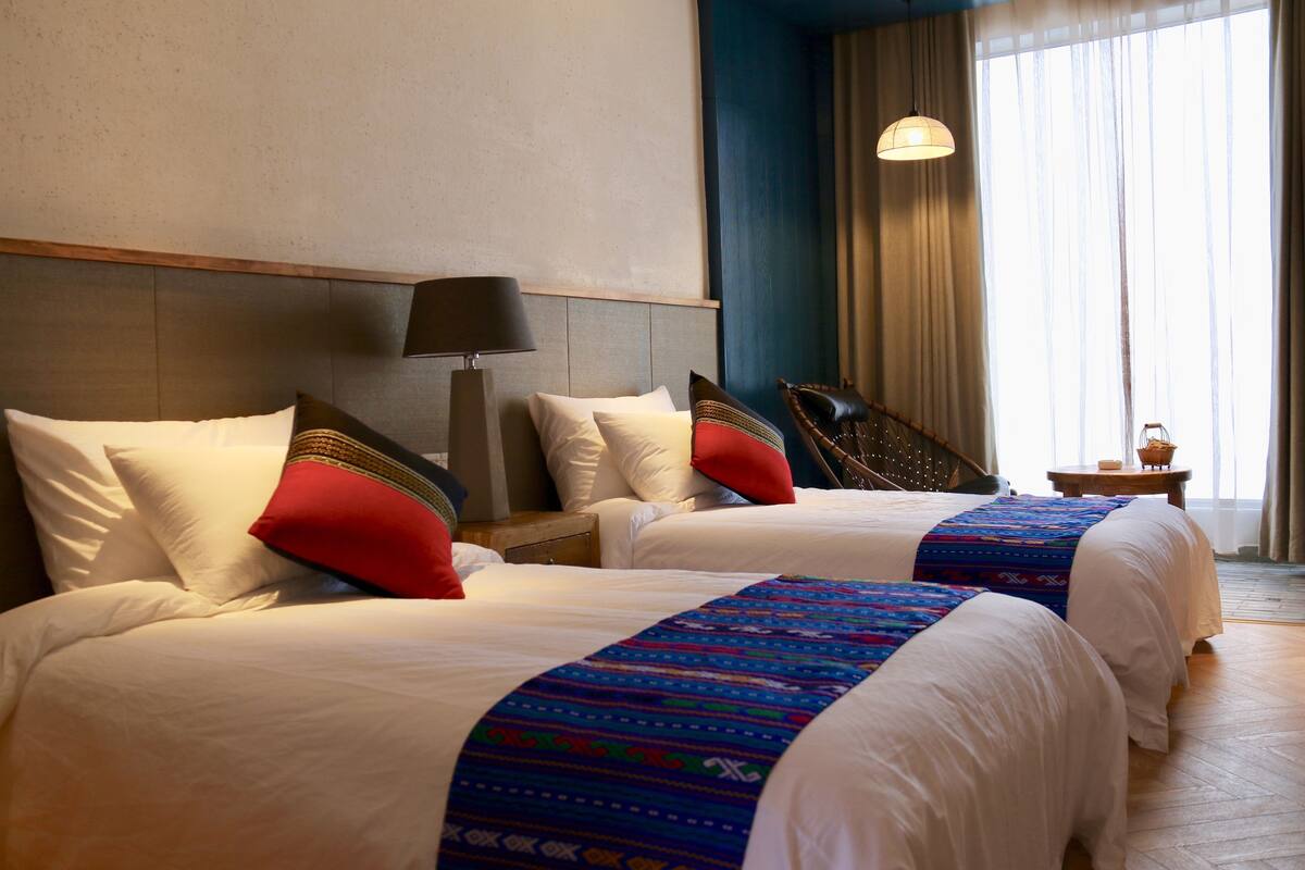 A hotel room with two beds, a nightstand, a lamp, a window with curtains, and a view of the outside.