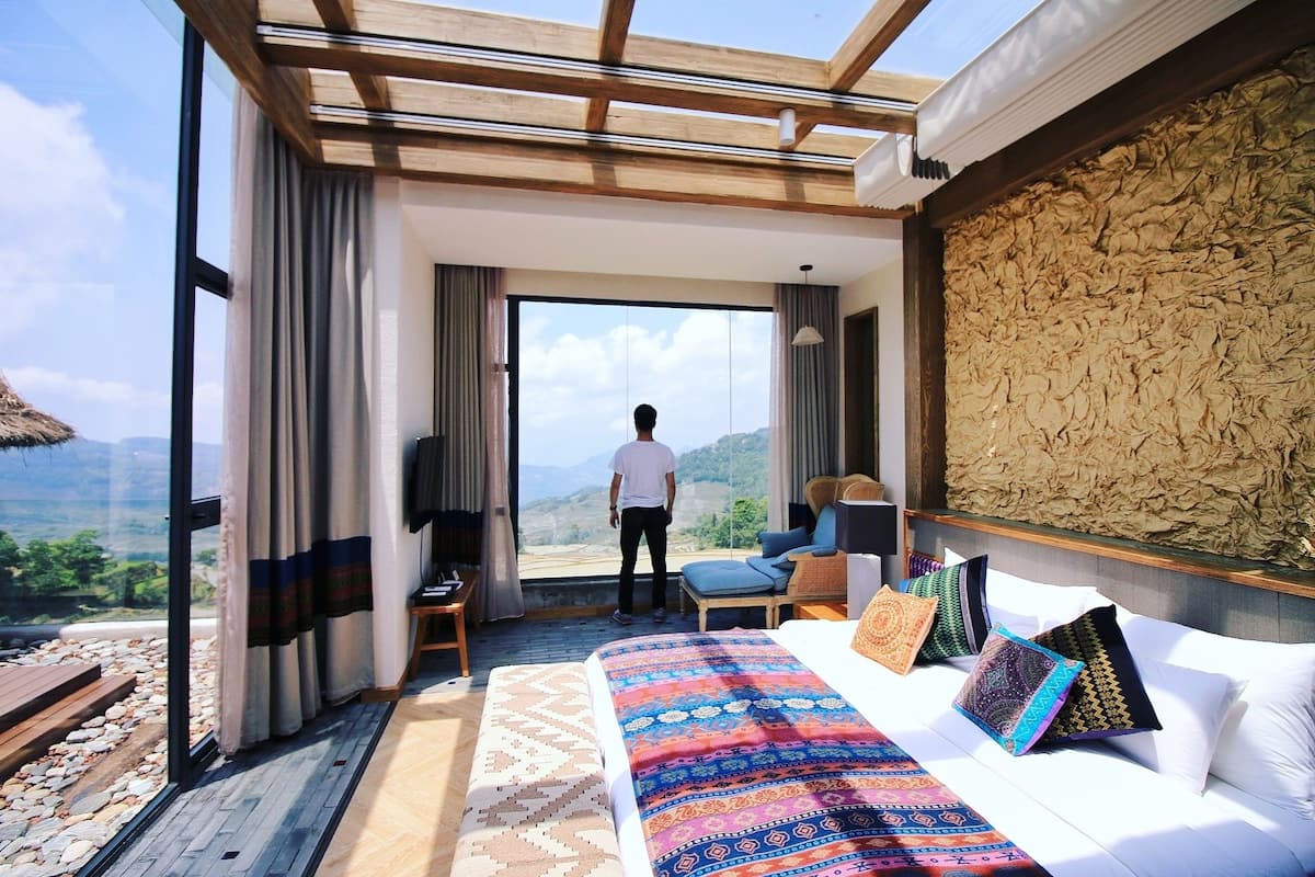 A bedroom with a large bed, a view of the outdoors, and a wooden ceiling structure.