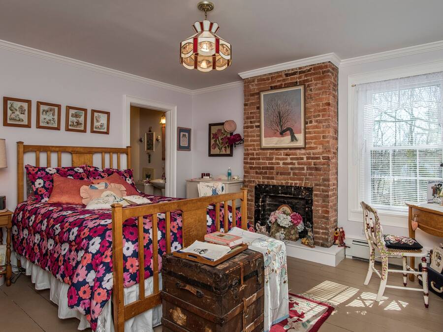 A cozy bedroom with a wooden bed, a brick fireplace, and a large window with blinds.