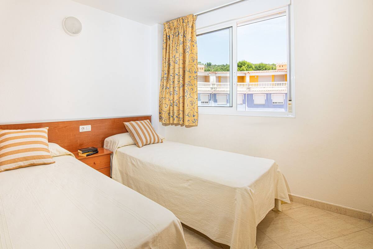 Habitació d'hotel amb dos llits individuals, un armari de fusta, una tauleta de nit amb un llibre i una finestra amb cortines.