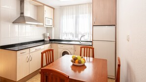 A compact kitchen with a dishwasher, a coffee maker, a microwave, and a refrigerator. There is a wooden dining table with a bowl of fruit. - Residencial Marcomar Casa Azahar (Alcala de Xivert)