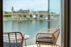 A balcony with wicker chairs and a table, overlooking a river with buildings in the background.