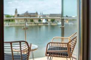 Un balcón con sillas de mimbre y una mesa, con vistas a un río y edificios al fondo.