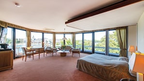 Habitación de hotel con una cama grande, un escritorio, dos sillas, una mesa de centro y vistas a la ciudad a través de grandes ventanales.