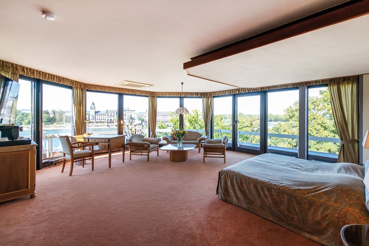 Habitación de hotel con una cama grande, un escritorio, dos sillas, una mesa de centro y vistas a la ciudad a través de grandes ventanales.