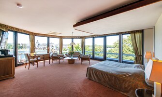 Habitación de hotel con una cama grande, un escritorio, dos sillas, una mesa de centro y vistas a la ciudad a través de grandes ventanales.