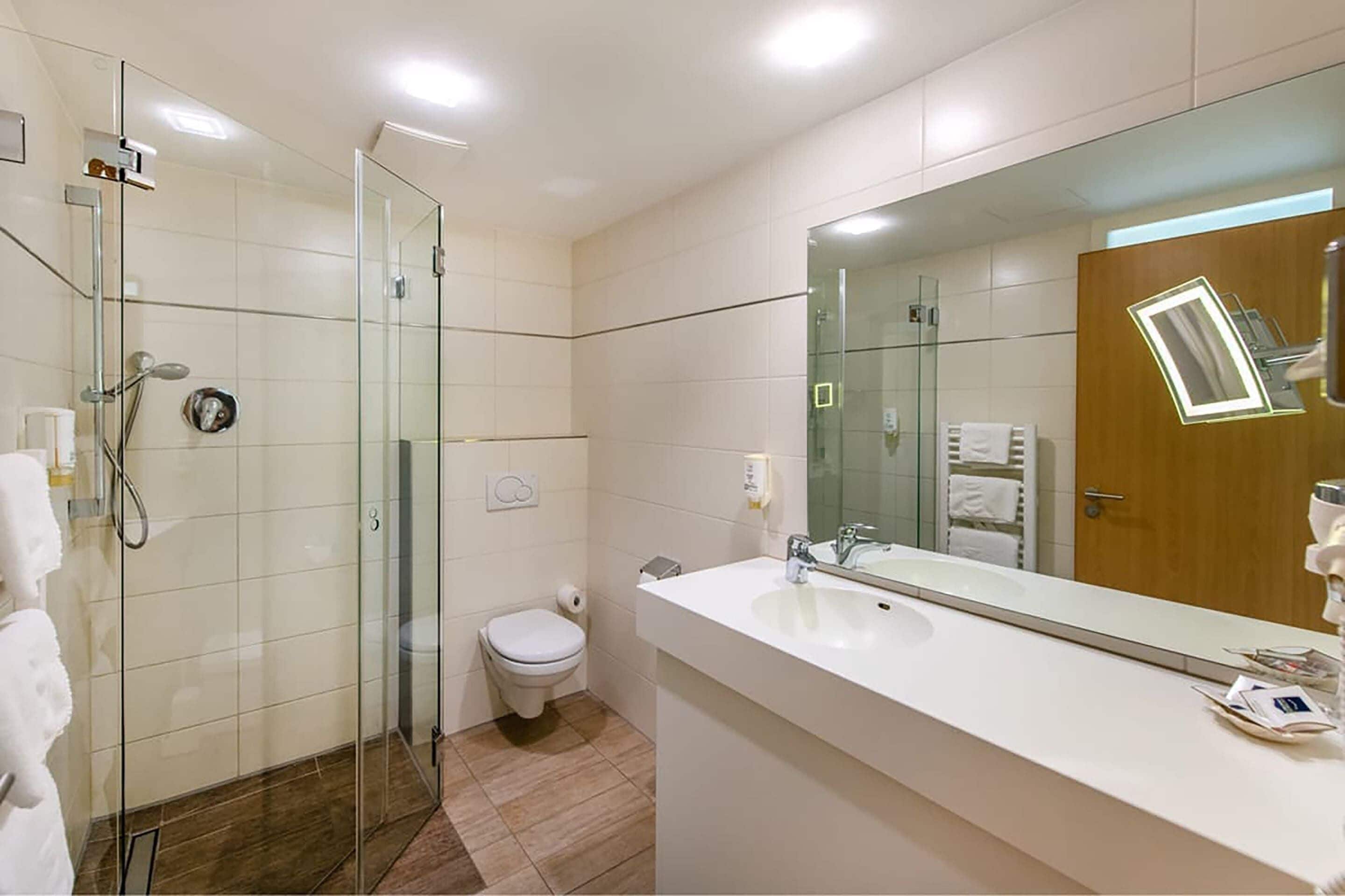 Ein modernes Badezimmer mit Duschkabine aus Glas, einer weißen Toilette und einem großen Spiegel über dem Waschbecken.