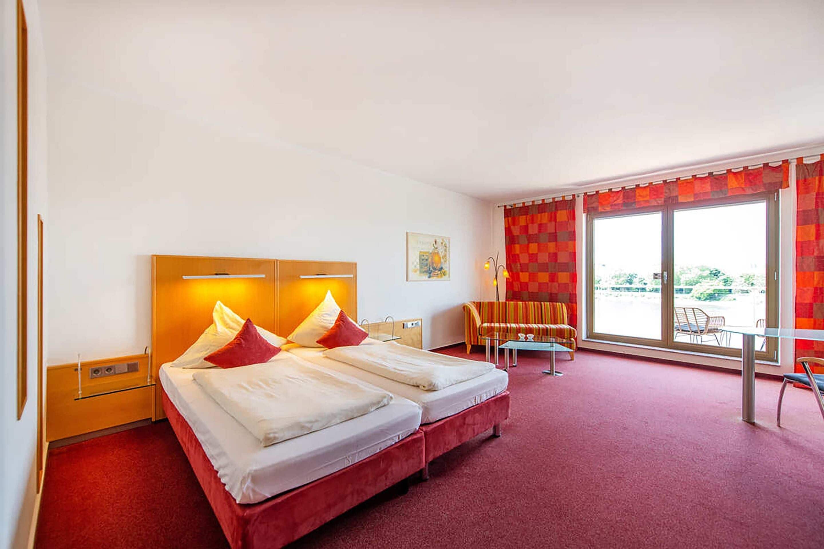 Habitación de hotel con dos camas, un sofá, una mesa de comedor y un amplio ventanal con cortinas.