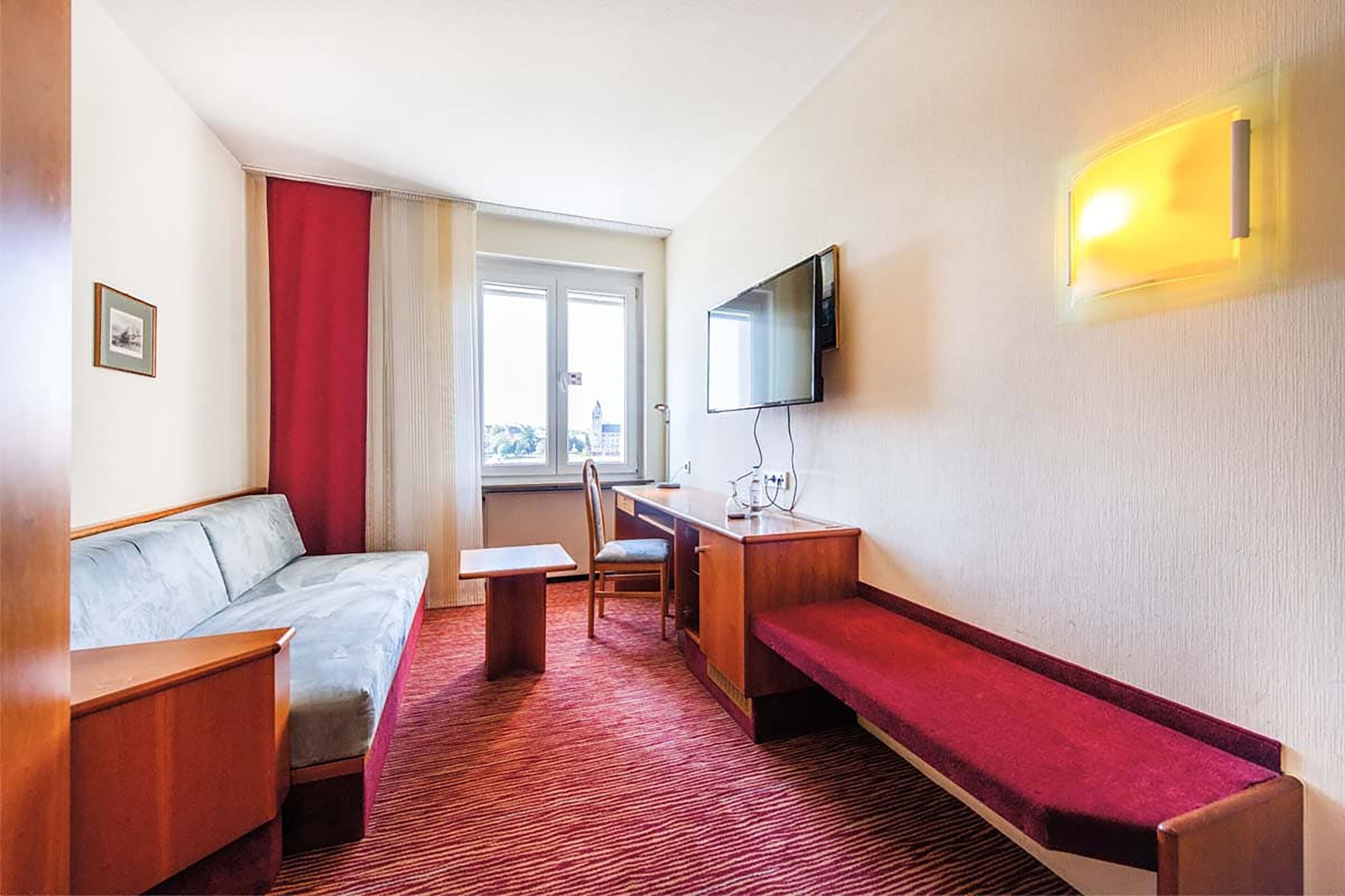 Une chambre d’hôtel de taille réduite, comprenant un canapé, un petit bureau, un banc, une télévision et une fenêtre avec des rideaux.