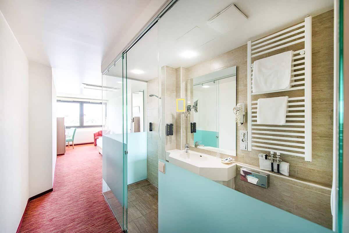 Un baño de hotel moderno con mampara de vidrio, un espejo grande, un perchero y un lavamanos.