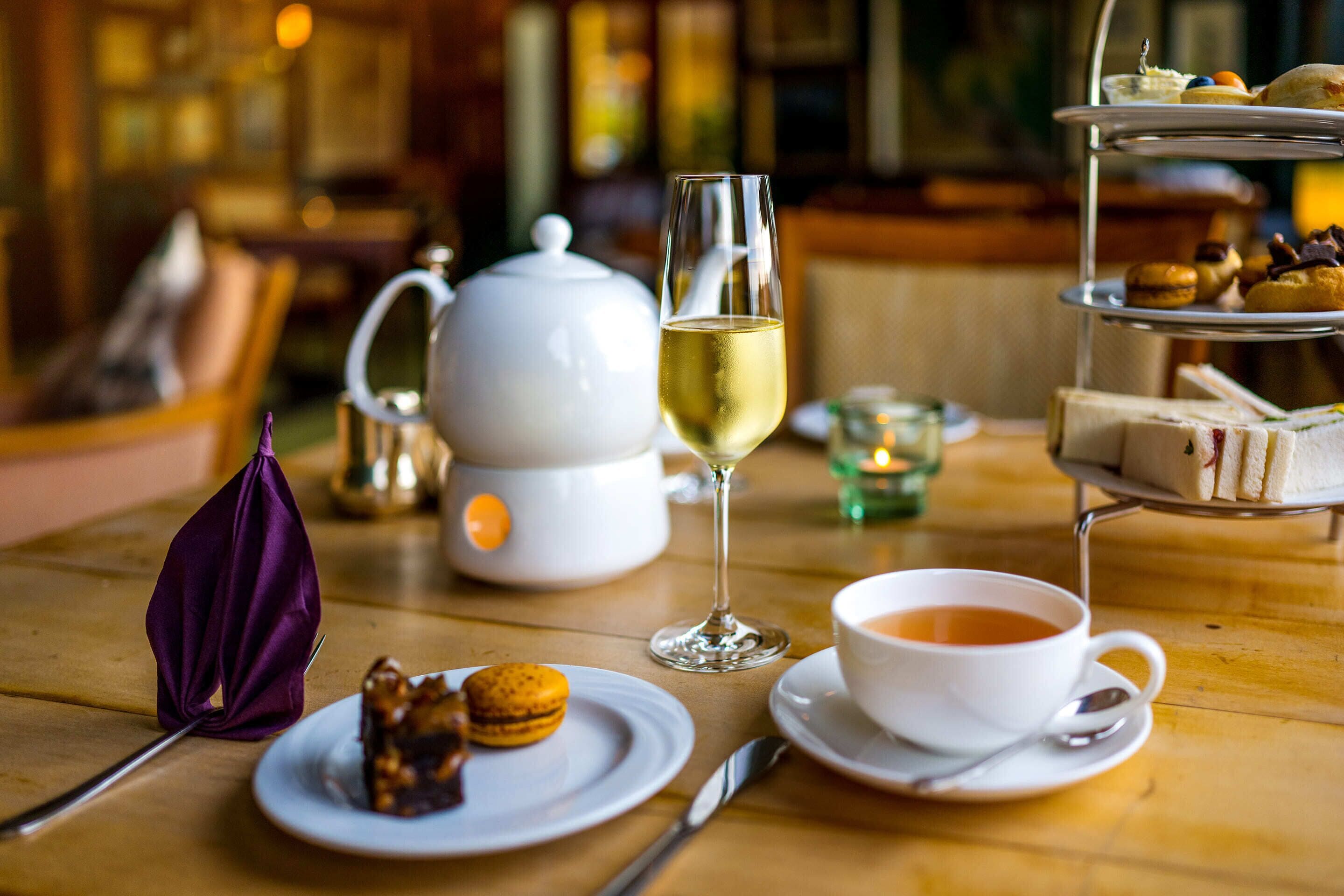Una mesa con un juego de té, una copa de vino blanco, una taza de té, un plato con macarons y un pastelillo con sándwiches.