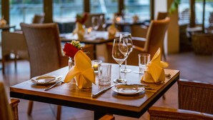 Una mesa de comedor dispuesta con platos, cubiertos, vasos y servilletas, con vistas al exterior a través de amplios ventanales.