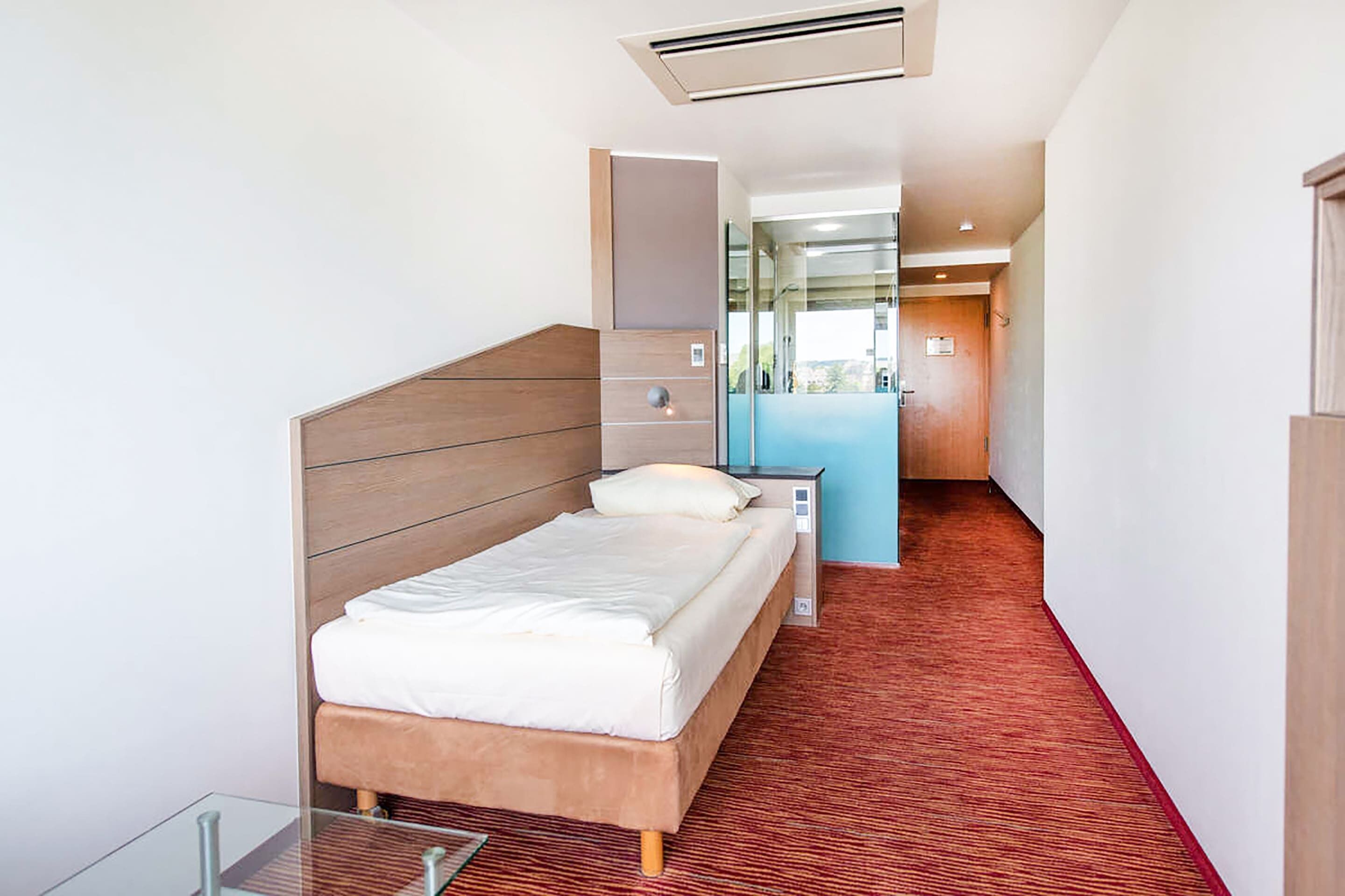 A modern hotel room with a wooden headboard bed, a glass table, and a red carpeted floor.