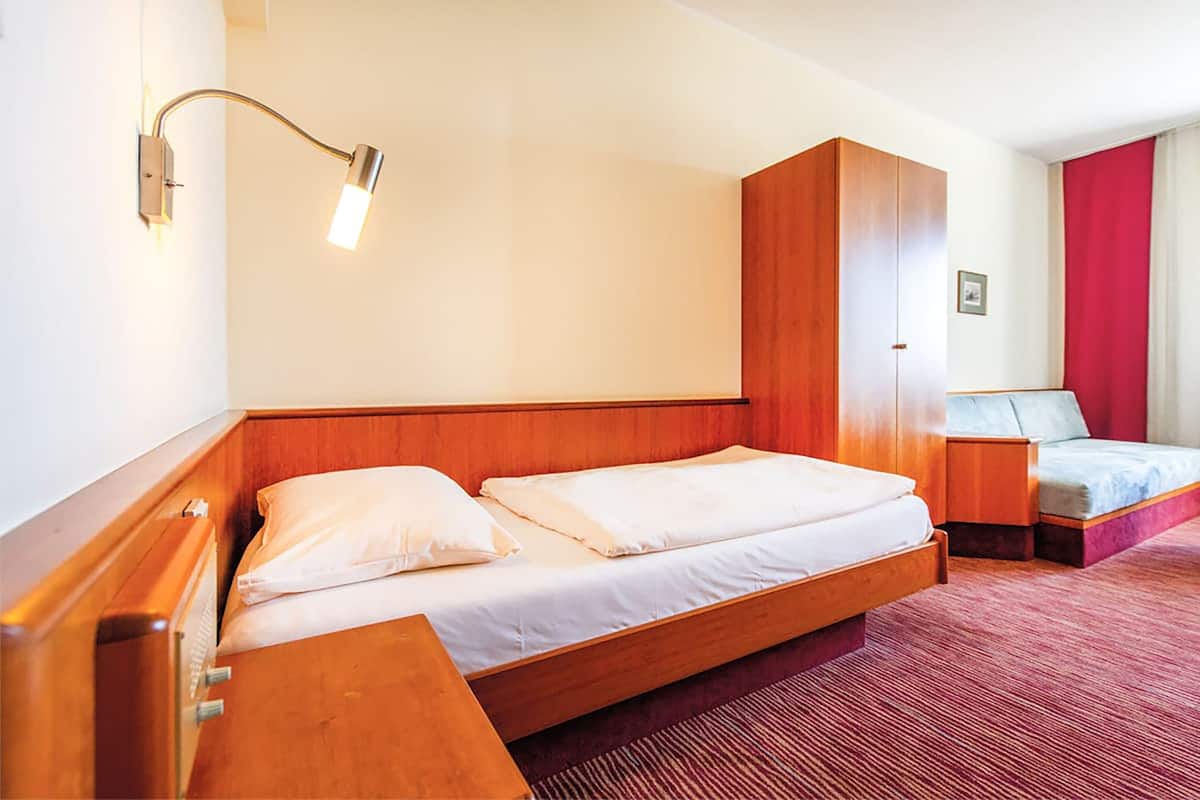 Habitación de hotel con cama de madera, mesita de noche, alfombra roja y lámpara de pared.