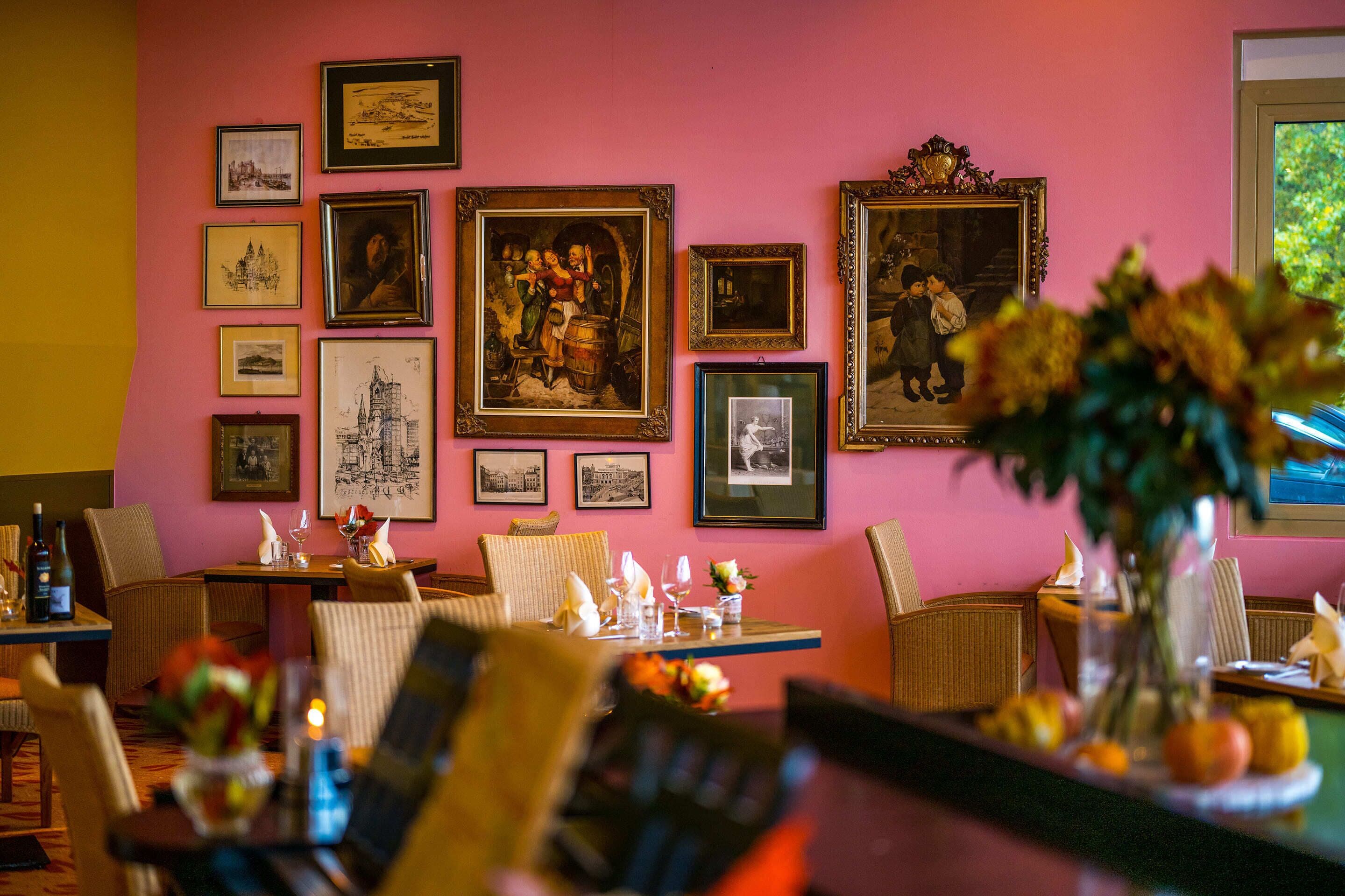 A dining area with a pink wall adorned with framed pictures, wicker chairs, and a table set for dining with a vase of flowers.