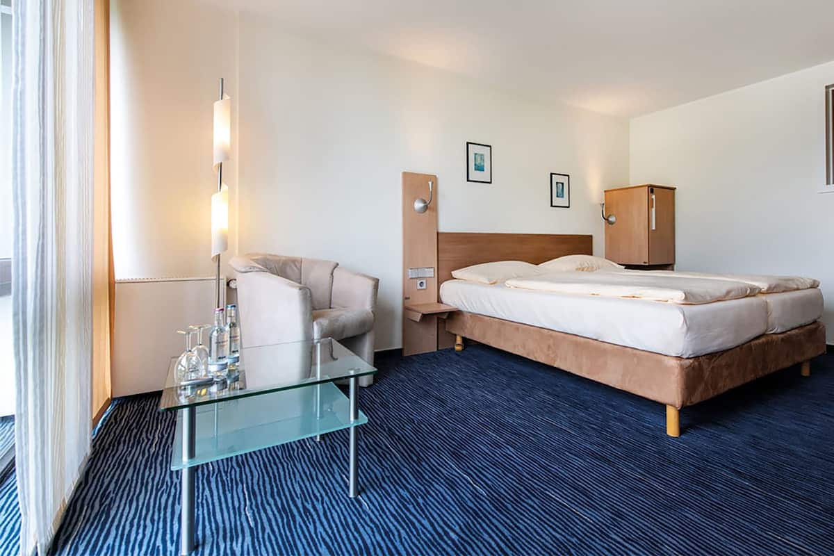 Una habitación de hotel con cama, mesa de vidrio, una silla, mesita de noche, armario y lámpara.