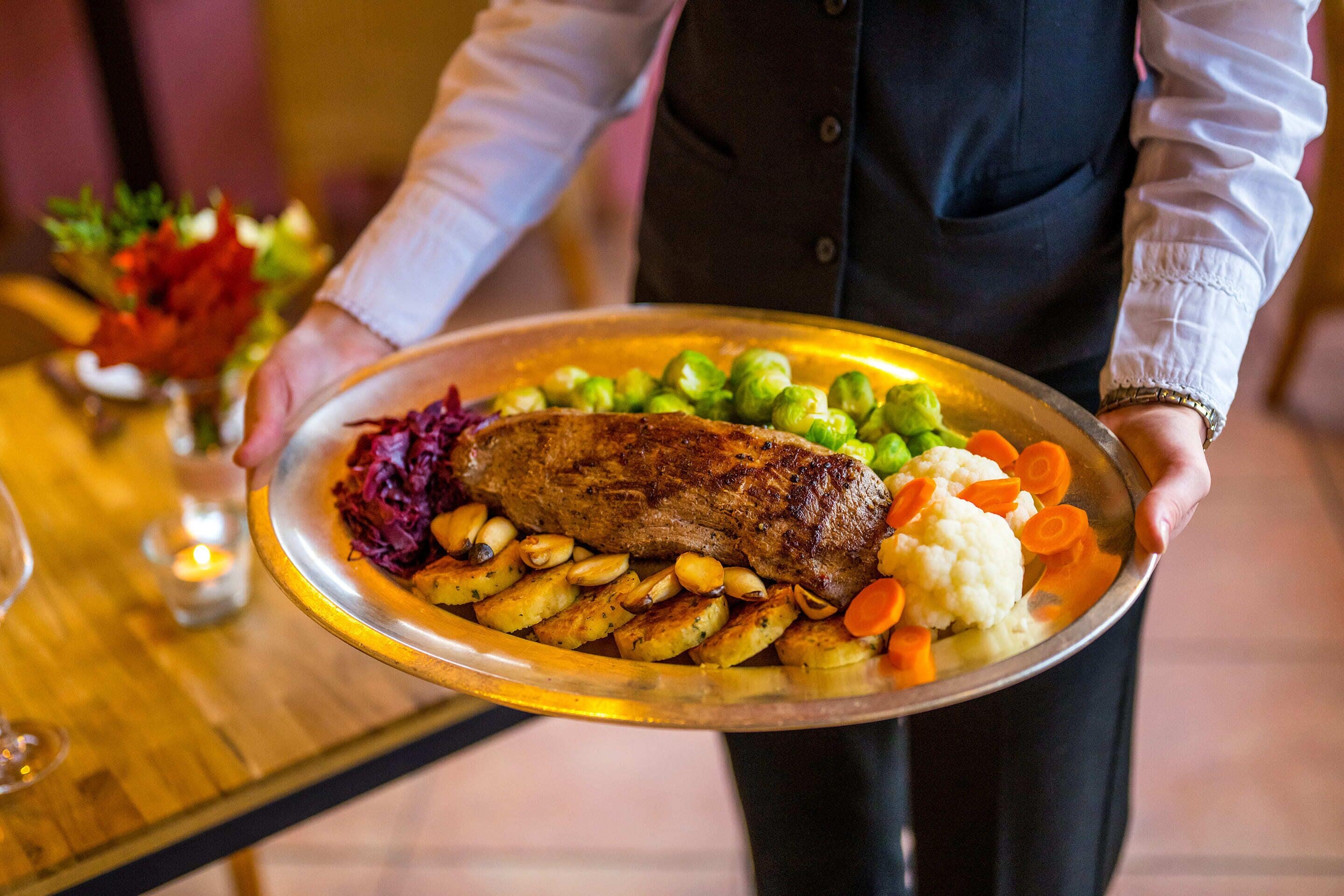 A person holding a large platter with roasted meat, potatoes, carrots, and Brussels sprouts.