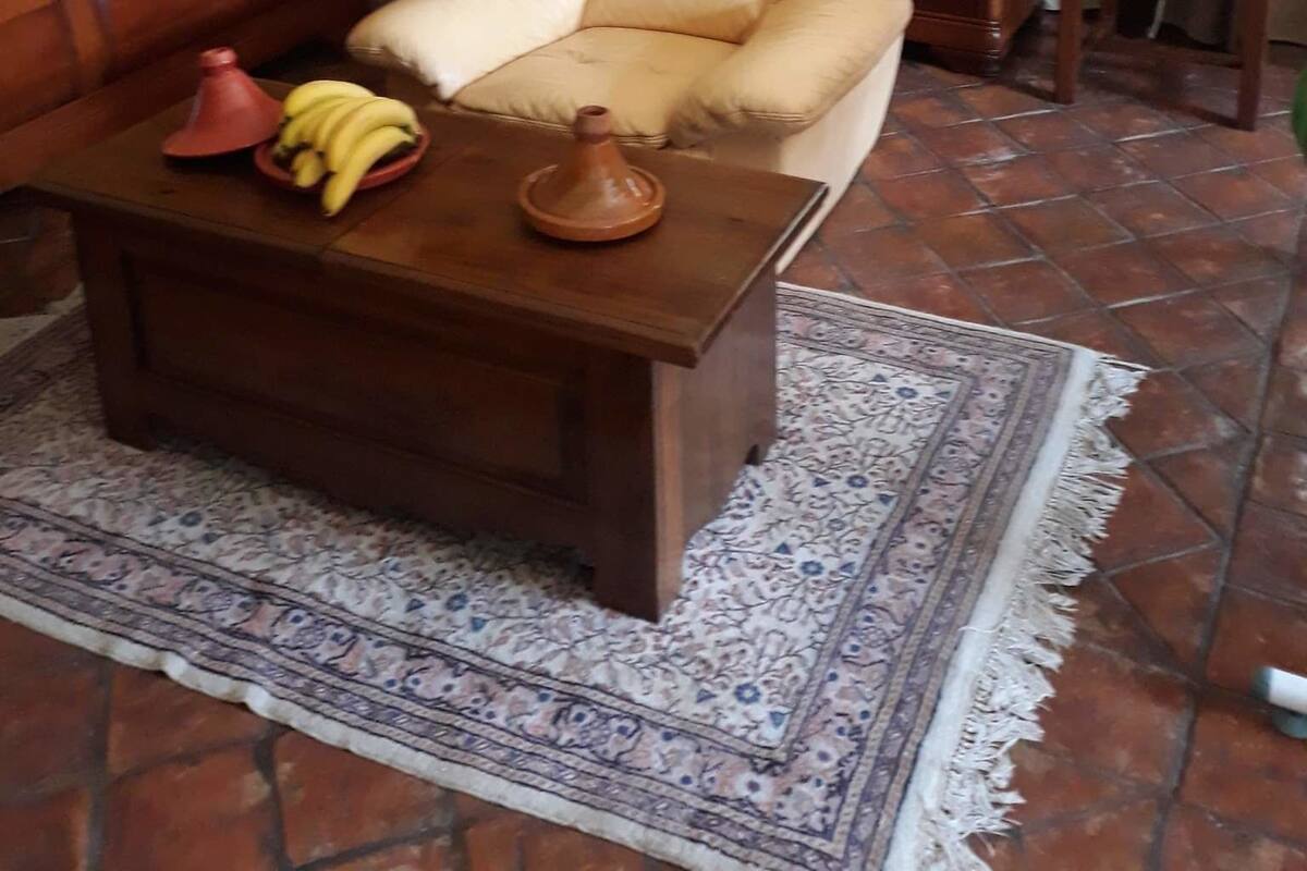 A living room with a leather armchair, a wooden coffee table with a red lamp, a patterned rug, and a bookshelf filled with books and framed pictures.