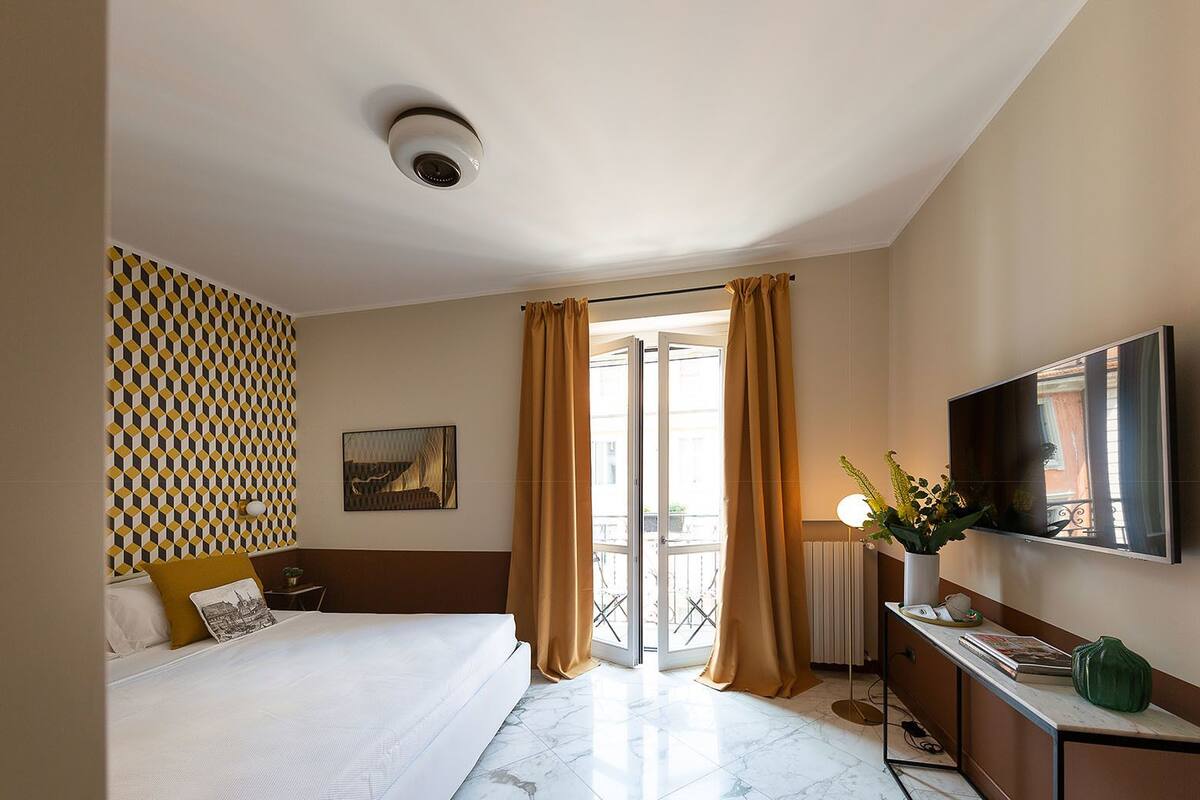 A hotel room with a bed, a TV, a desk, and a balcony with curtains.