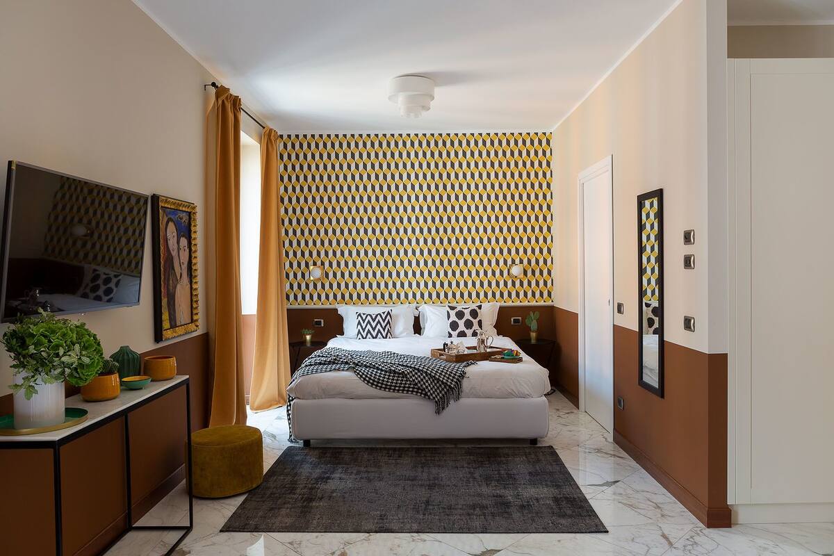 A modern bedroom with a patterned wallpaper, a bed with multiple pillows, a bedside table with a plant, and a mirror on the wall.