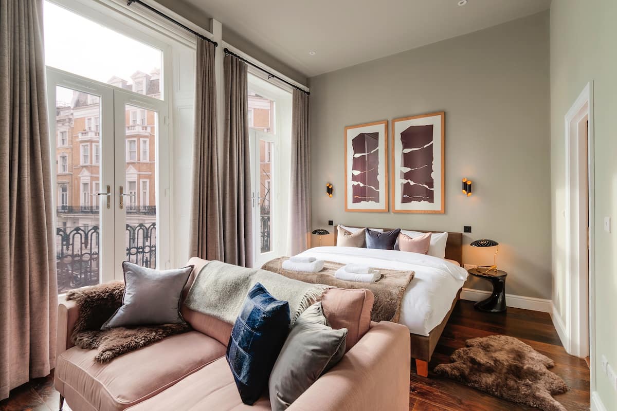 Una habitación de hotel con una cama grande, un sofá, una ventana con cortinas y dos cuadros en la pared.