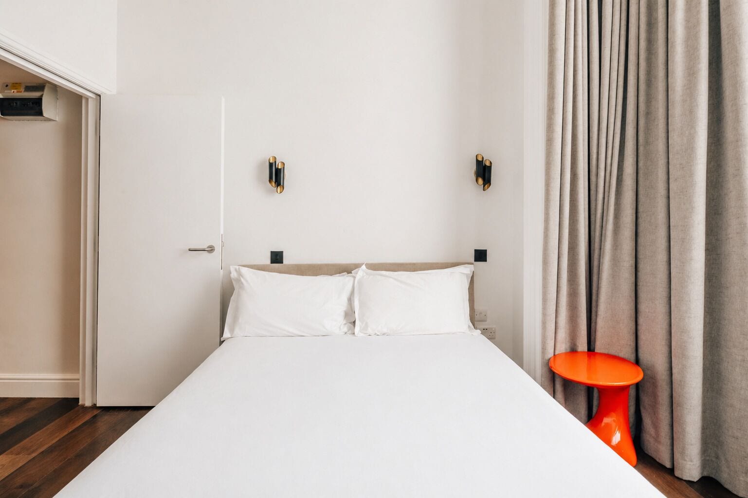 A hotel room with a bed, a door, a wall-mounted light fixture, and a curtain.