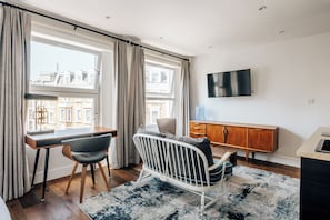 Ett modernt rum med ett skrivbord, en stol, en spjälsäng, en tv och utsikt över byggnader.