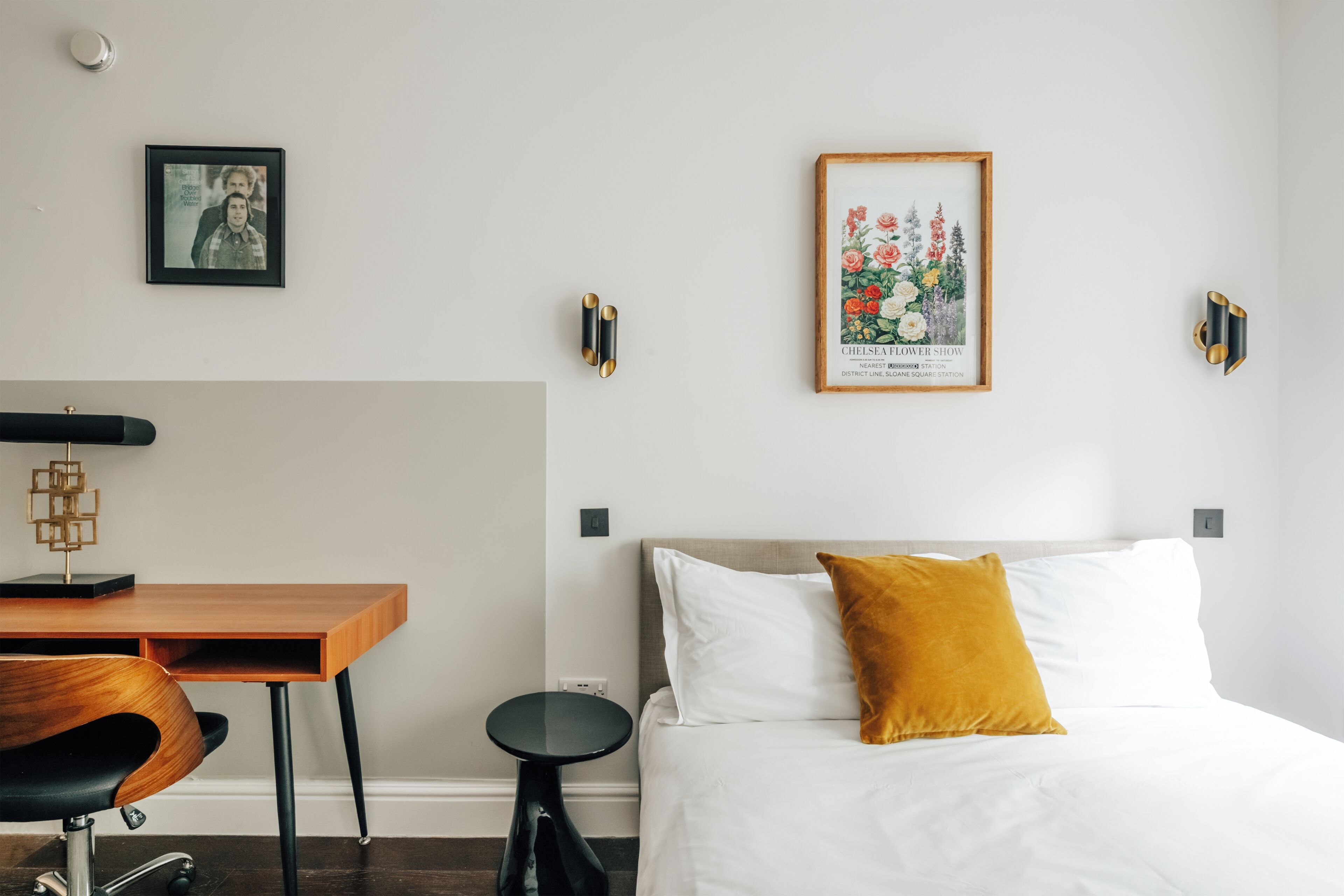 Quarto de hotel com uma cama, uma escrivaninha, uma cadeira, uma mesinha e duas luminárias fixadas na parede.
