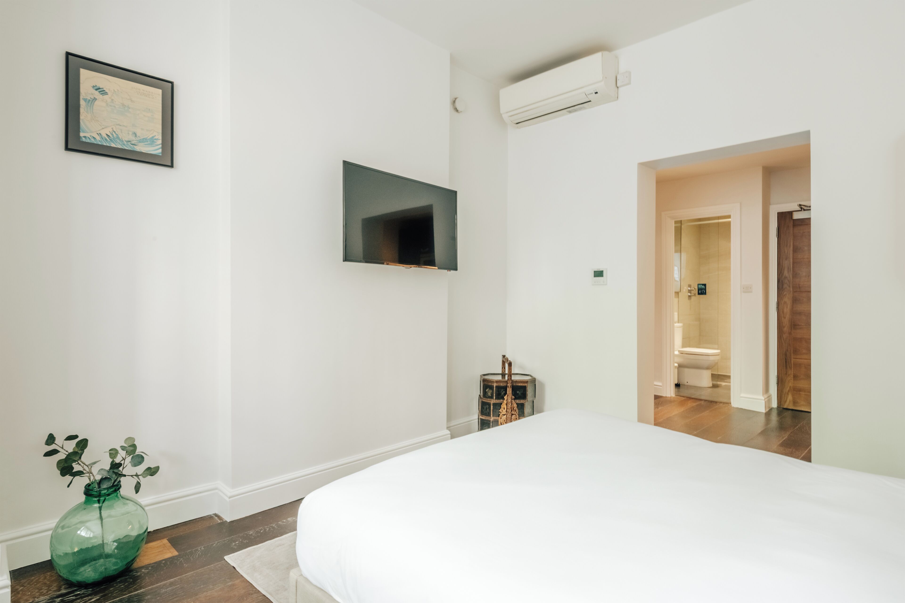 Een slaapkamer met een bed, een aan de muur bevestigde televisie, een groene vaas met planten, en een houten deur die naar een badkamer leidt.