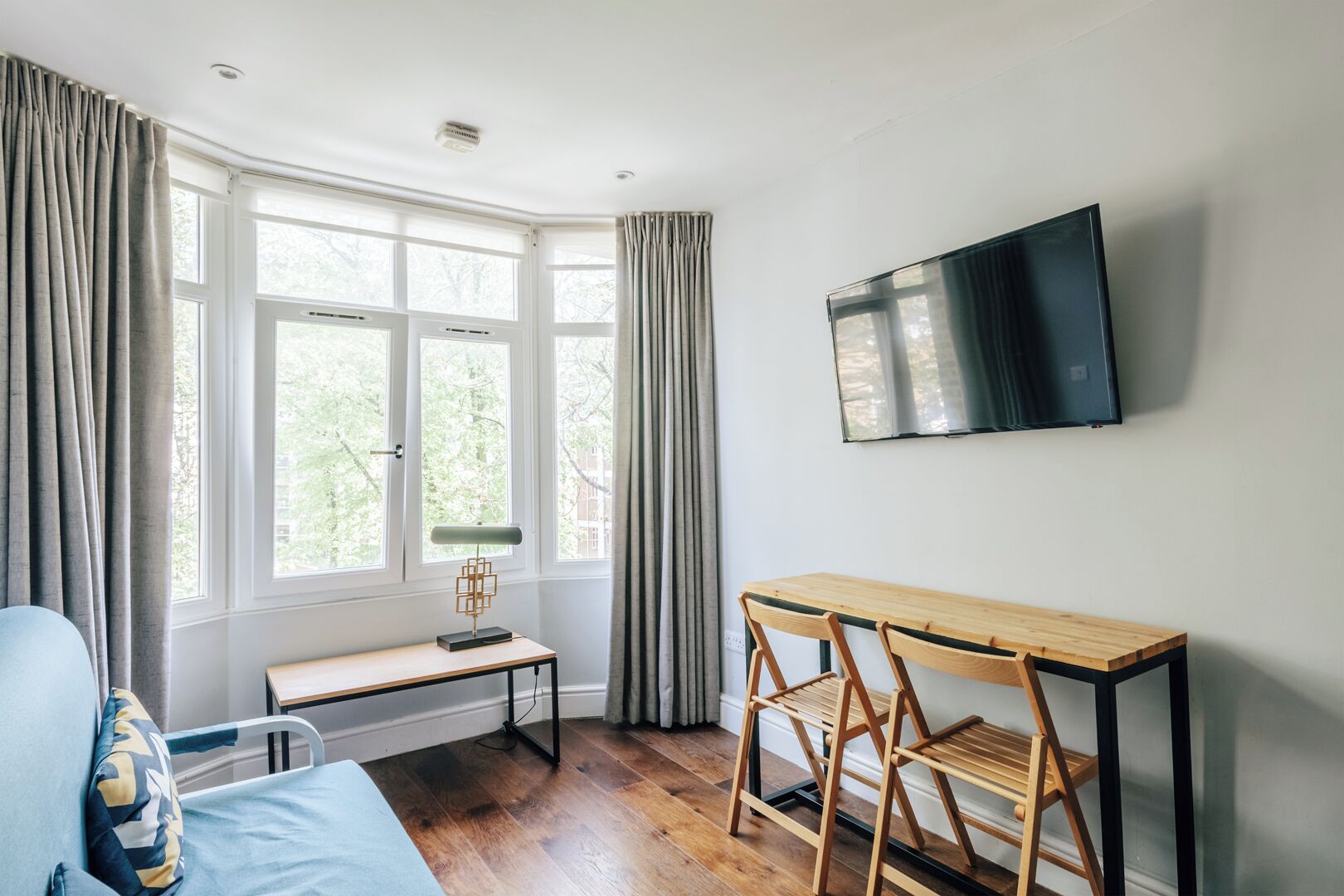 A room with a bed, a desk, a chair, a TV, and large windows with curtains.