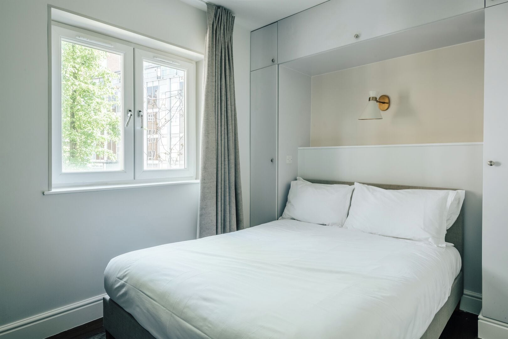 Un dormitorio con una cama, una ventana con cortinas, una lámpara de pared y vistas a árboles fuera.