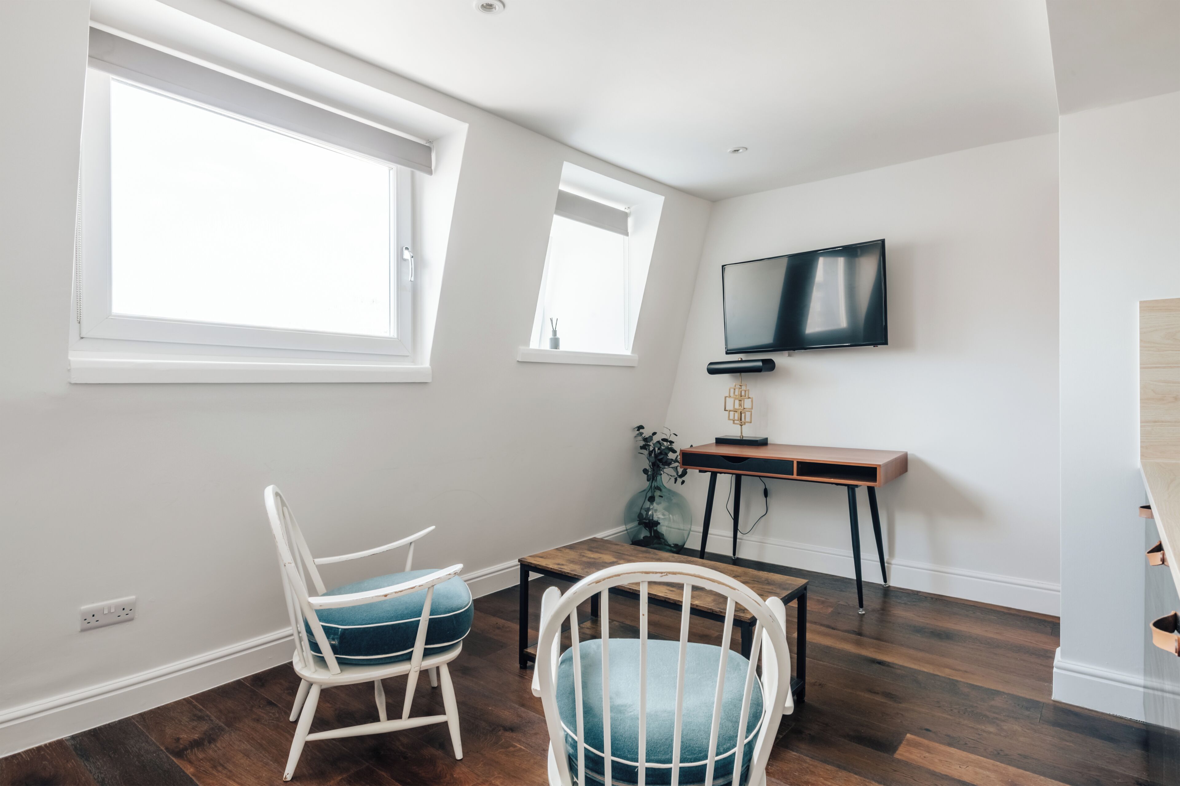 Ein Zimmer mit großem Fenster, einem an der Wand befestigten Fernseher, einem Holztisch und einem weißen Stuhl.