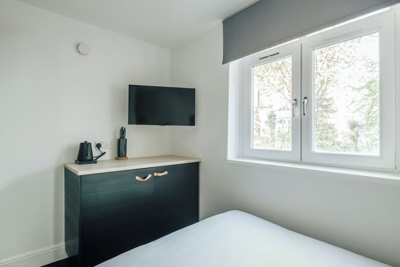 A bedroom with a bed, a bedside table with a kettle, a TV mounted above the table, and a window with a view of trees.