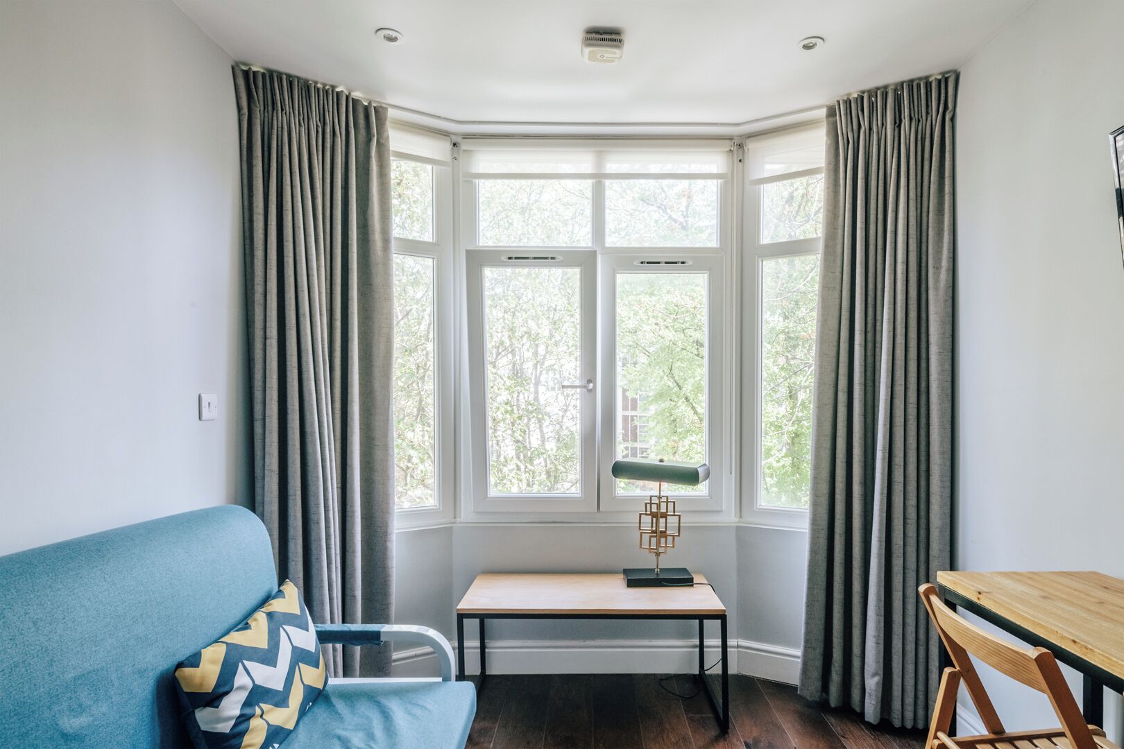 A room with a bay window, a blue sofa, a small table with a lamp, and a wooden chair.