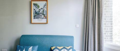Una habitación con un sofá azul, dos almohadones estampados, un cuadro de Chelsea, Londres, y una ventana con cortinas.