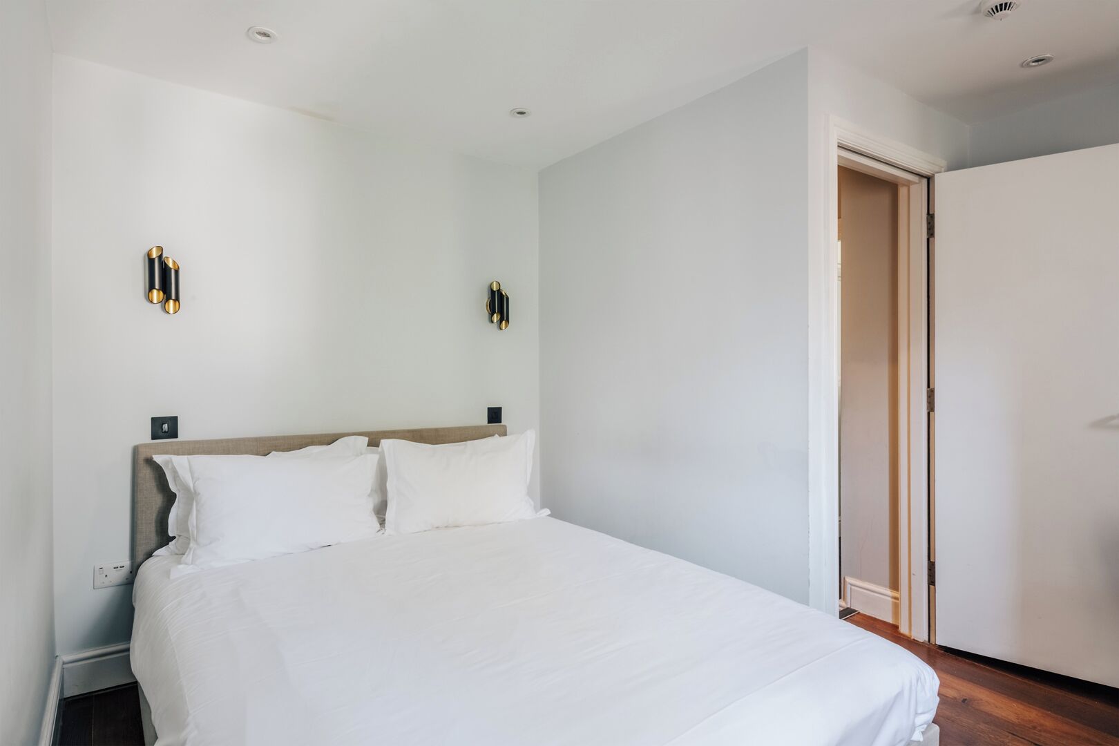 A bedroom with a large bed, a wooden headboard, two wall-mounted light fixtures, and a door to the right.