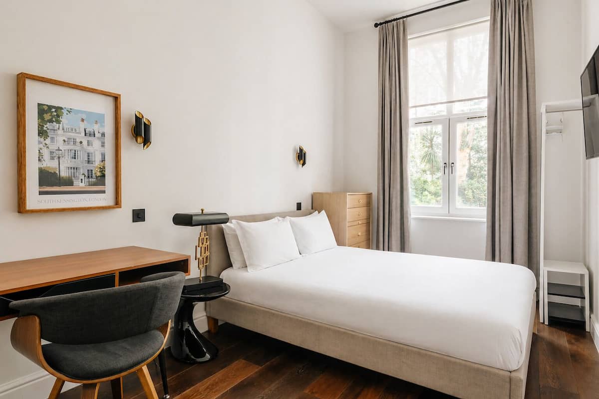 Habitación de hotel con cama, escritorio, silla, lámpara, cómoda y ventana con cortinas.