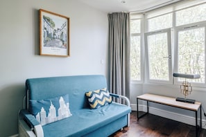 Ett rum med en blå soffa, en inramad bild av Chelsea i London, ett fönster med gardiner och ett skrivbord med en lampa.